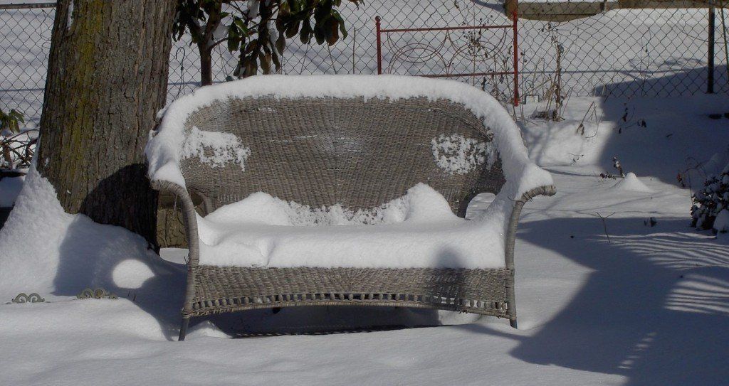 Snow-covered bench in a snowy yard beside a tree and chain-link fence.